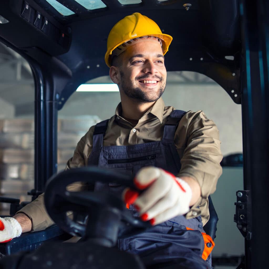 Male in working uniform and yellow hardhat operating forklift machine in warehouse storage room